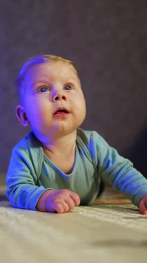 Sevimli sarışın bebek karnının üstünde uzanmış merakla yukarı bakıyor. Sevimli çocuk evde yerde, oyuncakların arasında yatıyor. Dikey video