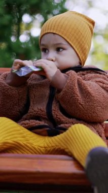 Güzel beyaz çocuk bir sürüden atıştırmalık yiyor. Çocuk parkta bankta oturuyor. Düşük açı. Dikey video