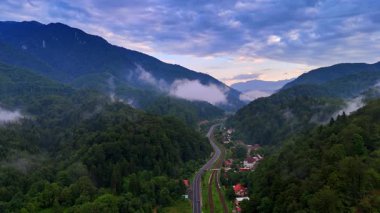 Ormanlık dağların arasındaki yerleşim bölgesinin görüntüleri. İnsansız hava aracı, tren yolu ve otoban üzerinden köyü geçiyor. Dramatik bulutlar manzaranın üzerindeki gökyüzünü kaplıyor. Romanya kırsal kesimi.