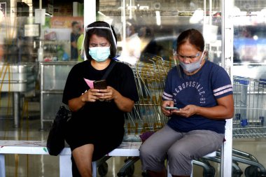 Antipolo City, Philippines - July 16, 2020: Filipina ladies wearing face shield and mask use their smartphone to pass the time while waiting outside a supermarket during the Covid 19 outbreak.