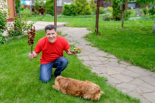 The senior man is sitting on the grass with shish kebab and a plate with vegetables in his hands and smiling. Near him lay his dog.