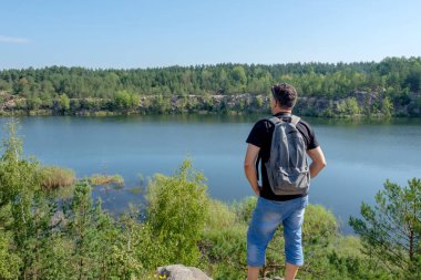 Sırt çantası ile yetişkin beyaz adam kayanın üzerinde duruyor ve gölde görünüyor.