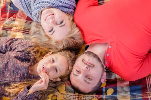 Young beautiful family lying on a plaid and smiling on a sunny autumn day. Little daughter eat sandwich. The concept of a happy family and family with one children.