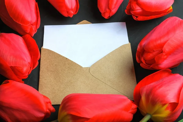 Mockup de ramo de tulipanes rojos frescos y tarjeta de felicitación vacía en sobre kraft sobre fondo oscuro con espacio para copiar . — Foto de Stock