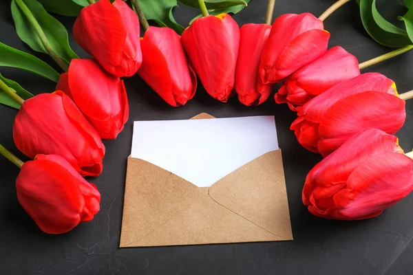 Mockup de ramo de tulipanes rojos frescos y tarjeta de felicitación en blanco en sobre kraft sobre fondo oscuro con espacio de copia . — Foto de Stock