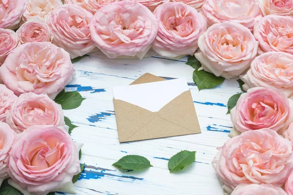 Composición de flores. Sobre de papel artesanal abierto y hojas en el marco de rosas rosadas en una mesa de madera envejecida . — Foto de Stock