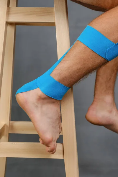 Close-up kinesiology tape applied to injured calf and heel of a white male sitting on a tabouret. Studio shot, vertical orientation