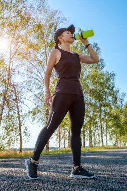 Dinlenen genç beyaz kadın kırsal yolda sıkı çalıştıktan sonra plastik şişeden su içiyor. Susuzluk, sıcaklık, dayanıklılık ve su dengesinin sağlanması. Dikey yönelim