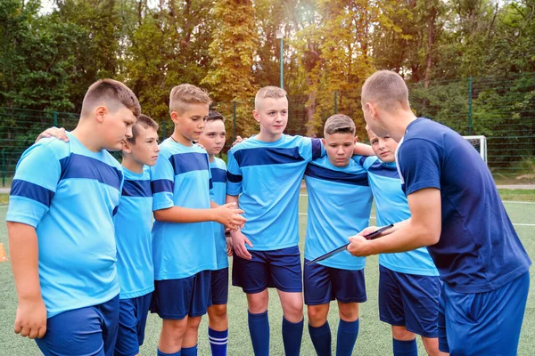 Děti ve fotbalové uniformě se objímají a poslouchají vysvětlování trenéra. — Stock fotografie