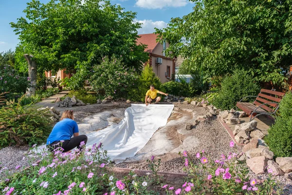 Family roll out a roll of white non-woven geotextile fabric on the ground before laying polyethylene film to set up a fish pond in backyard near their home.