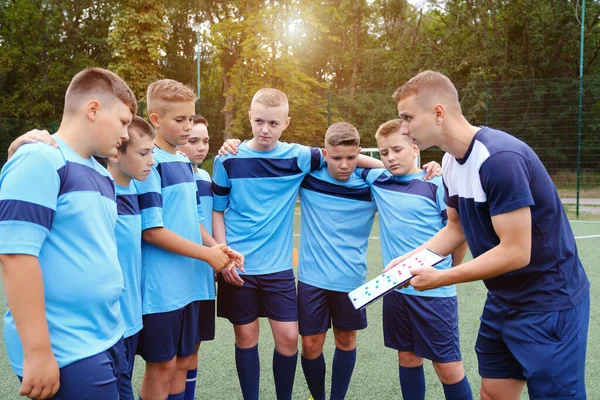 Children in football uniform hugs and listening to explain of coach with clipboard in hands. Young coach explaining football game strategy to kids soccer team.