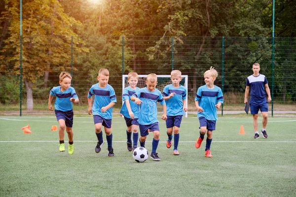 Çocuklar futbol antrenmanından önce ısınmak için futbol topunu tekmeliyor. Arka plandaki koç eğitim sürecini izliyor
