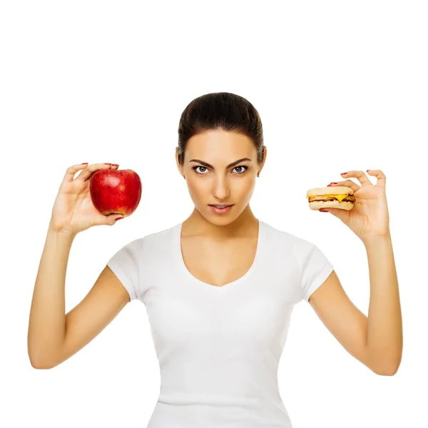 Die schöne junge brünette Frau im weißen T-Shirt hält Apfel und Hamburger in der Hand. steht lächelnd auf weißem Hintergrund — Stockfoto