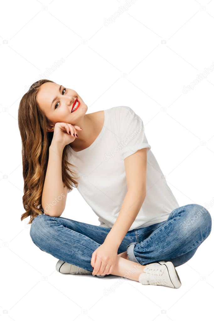 young cheerful girl in jeans, white t-shirt, sneakers sits on floor and smiles, tilting her head to side. isolated on white background