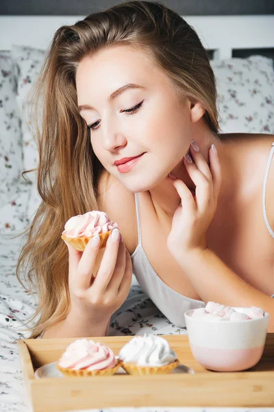 Jong blond meisje liggend in bed cupcake houdt in haar hand en zorgvuldig te kijken naar hem — Stockfoto