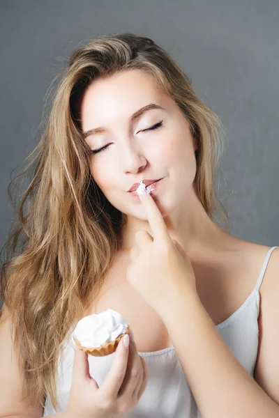 Joven hermosa rubia con el pelo largo sosteniendo una magdalena en la mano, disfrutando con los ojos cerrados. sobre fondo gris —  Fotos de Stock