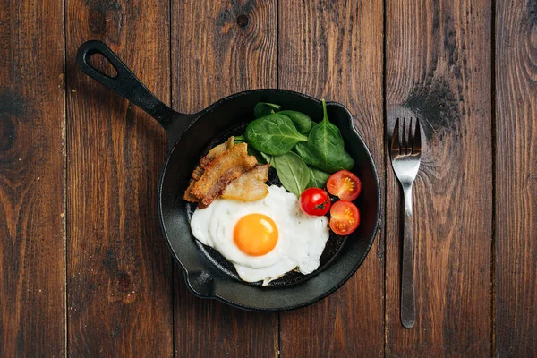 Ahşap masada kızartma tavası ile lezzetli pastırma, yumurta, ıspanak ve domates üst görünümü.