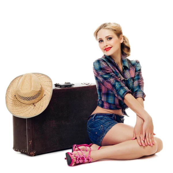 Chica rubia en camisa a cuadros y pantalones cortos de mezclilla está sentado cerca de una vieja maleta con sombrero de paja y sonriendo juguetonamente a la cámara. aislado sobre fondo blanco —  Fotos de Stock