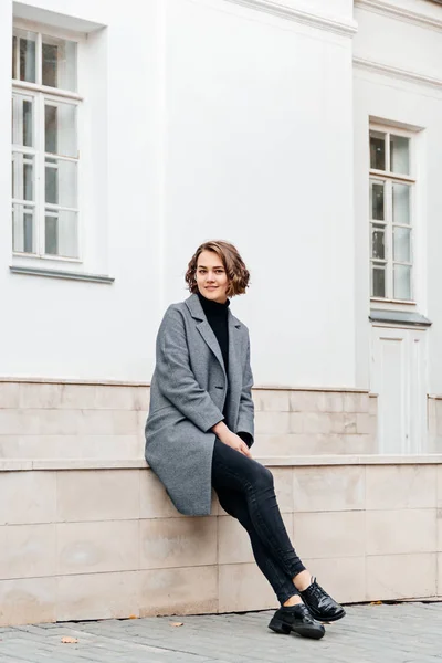 Meisje in grijze jas en zwart jeans zit in de buurt gebouw — Stockfoto