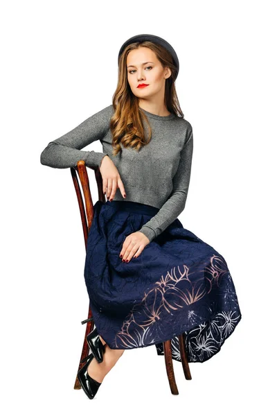 Young beautiful girl in dark blue skirt and gray jumper, and gray hat sitting on old chair. isolated on white background — Stock Photo, Image