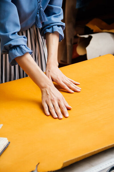 Young woman wearing casual clothes standing at table in her workshop rolling out piece of yellow leather material