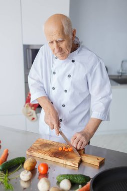 Gülümseyen bir ışık mutfakta bıçakla adam yaşlı. Büyükbaba gri saç ile bir akşam yemeği için sebzeleri Dilimleme.