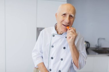Gri saçlı yaşlı Şef mutfakta bir havuç dilim tatma. Bir havuç ile mutlu bir adam.