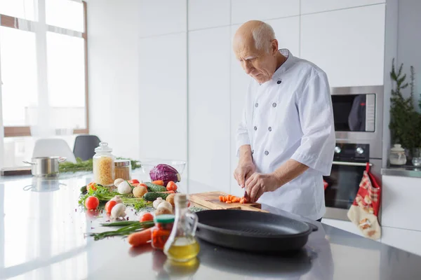 Happy elderly man is cooking dinner in the kitchen. A man with a knife is slicing vegetables for a salad. The concept of cooking.