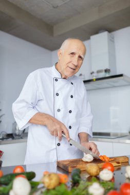 Gülümseyen bir ışık mutfakta bıçakla adam yaşlı. Büyükbaba gri saç ile bir akşam yemeği için sebzeleri Dilimleme.