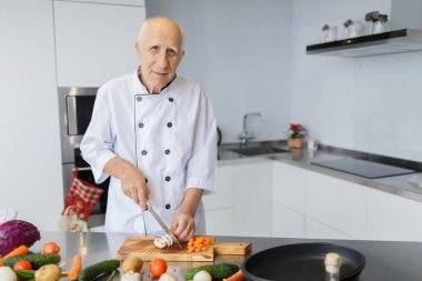 Gülümseyen bir ışık mutfakta bıçakla adam yaşlı. Büyükbaba gri saç ile bir akşam yemeği için sebzeleri Dilimleme.