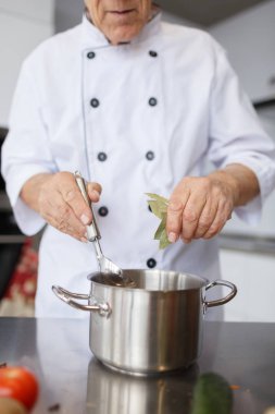 Yaşlı Şef bir çorba yemek pişiriyor. Dedesi bir metal kaşık ve bir defne yaprağı çorbanın içinde mutfak ekleme pan ile.