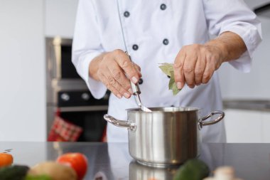 Yaşlı Şef bir çorba yemek pişiriyor. Dedesi bir metal kaşık ve bir defne yaprağı çorbanın içinde mutfak ekleme pan ile.