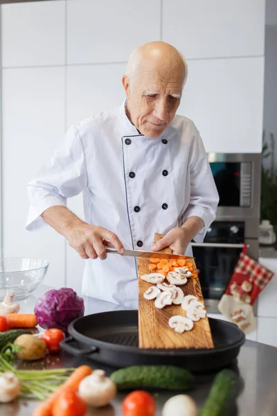 Happy elderly man is cooking dinner in the kitchen. Grandad with a knife is slicing vegetables for a salad. The concept of cooking.