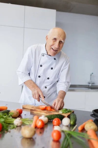 Happy elderly man is cooking dinner in the kitchen. Grandad with a knife is slicing vegetables for a salad. The concept of cooking.
