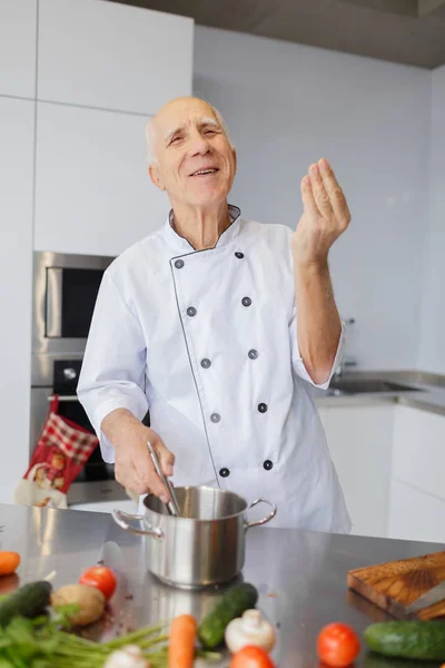 The chef is cooking a soup. The elderly man with a metal spoon and pan in the kitchen. Vegetarian soup