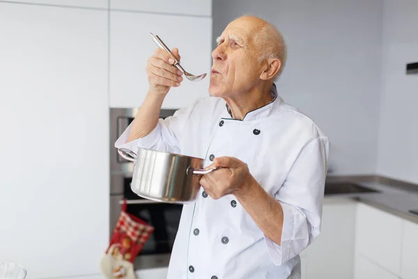 The elderly man with gray hair is tasting a soup which cooked before. Grandfather with a metal spoon and pan in the kitchen