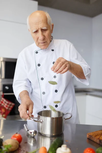 The elderly chef is cooking a soup. Grandfather with a metal spoon and pan adding a laurel leaf to the soup in the kitchen.