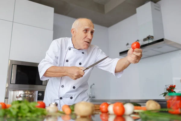 A grumpy aged man chef with a knife cutting tomatoes in the light kitchen. A concept of slicing vegetables for a dinner.