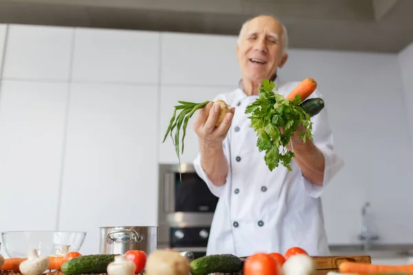 A happy aged man with vegetables for a salad in the kitchen. Grandad with grey hair holding vegetables for a dinner.