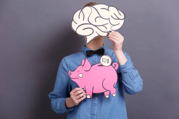 A young woman holds a big paper pink piggy bank with a coin and a big brain on a gray background. The concept of money and finance