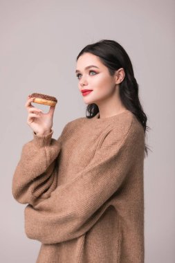 Kırmızı dudaklar çörek ile poz güzel kızla. Uzun saçlı, parlak kırmızı dudaklar, holding lezzetli çörek el ile çok seksi esmer kız kameraya bakıyor. Koyu gri arka plan, kopya alanı