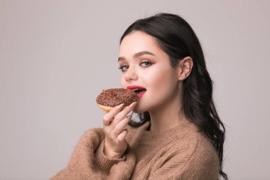 Kırmızı dudaklar çörek ile poz güzel kızla. Uzun saçlı, parlak kırmızı dudaklar, holding lezzetli çörek el ile çok seksi esmer kız kameraya bakıyor. Koyu gri arka plan, kopya alanı