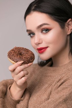 Kırmızı dudaklar çörek ile poz güzel kızla. Uzun saçlı, parlak kırmızı dudaklar, holding lezzetli çörek el ile çok seksi esmer kız kameraya bakıyor. Koyu gri arka plan, kopya alanı