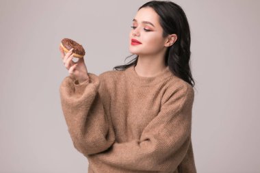 Kırmızı dudaklar çörek ile poz güzel kızla. Uzun saçlı, parlak kırmızı dudaklar, holding lezzetli çörek el ile çok seksi esmer kız kameraya bakıyor. Koyu gri arka plan, kopya alanı