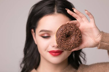 Kırmızı dudaklar çörek ile poz güzel kızla. Uzun saçlı, parlak kırmızı dudaklar, holding lezzetli çörek el ile çok seksi esmer kız kameraya bakıyor. Koyu gri arka plan, kopya alanı