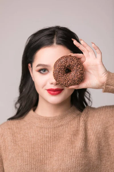 Kırmızı dudaklar çörek ile poz güzel kızla. Uzun saçlı, parlak kırmızı dudaklar, holding lezzetli çörek el ile çok seksi esmer kız kameraya bakıyor. Koyu gri arka plan, kopya alanı