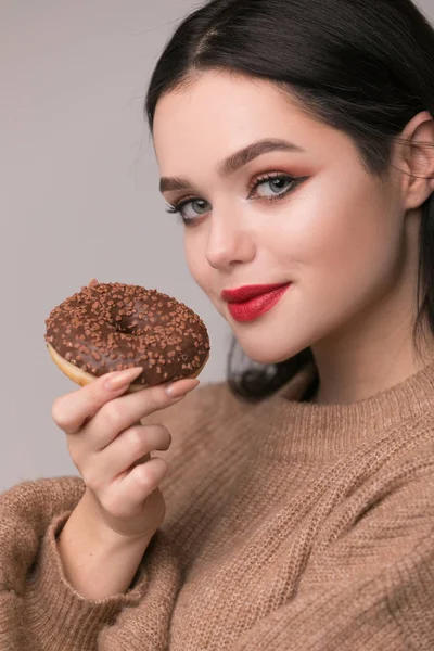 Kırmızı dudaklar çörek ile poz güzel kızla. Uzun saçlı, parlak kırmızı dudaklar, holding lezzetli çörek el ile çok seksi esmer kız kameraya bakıyor. Koyu gri arka plan, kopya alanı