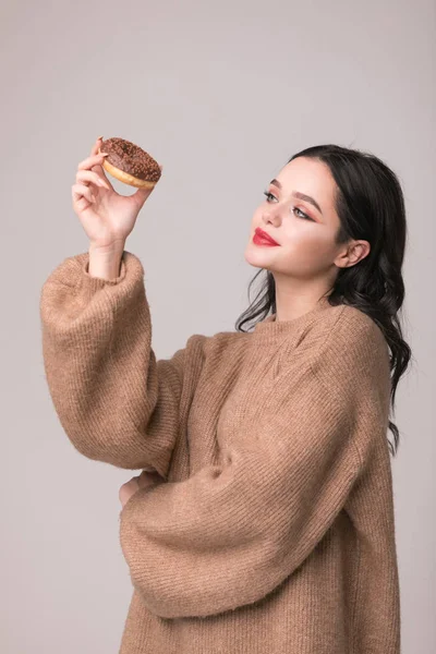 Kırmızı dudaklar çörek ile poz güzel kızla. Uzun saçlı, parlak kırmızı dudaklar, holding lezzetli çörek el ile çok seksi esmer kız kameraya bakıyor. Koyu gri arka plan, kopya alanı