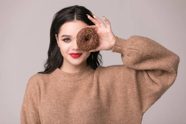 Kırmızı dudaklar çörek ile poz güzel kızla. Uzun saçlı, parlak kırmızı dudaklar, holding lezzetli çörek el ile çok seksi esmer kız kameraya bakıyor. Koyu gri arka plan, kopya alanı