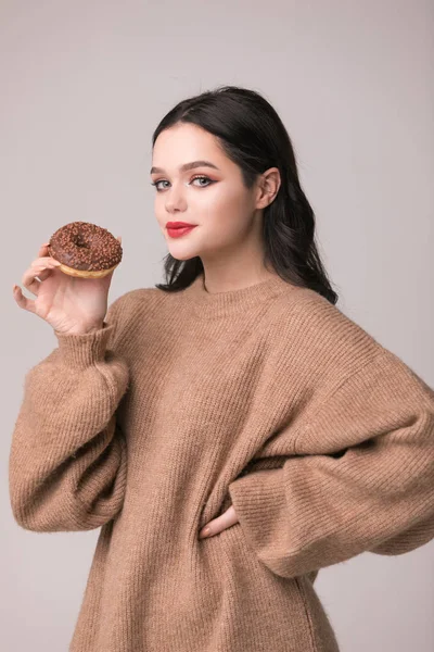 Kırmızı dudaklar çörek ile poz güzel kızla. Uzun saçlı, parlak kırmızı dudaklar, holding lezzetli çörek el ile çok seksi esmer kız kameraya bakıyor. Koyu gri arka plan, kopya alanı